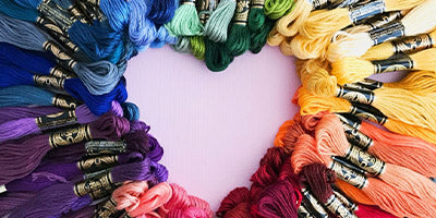 Embroidery threads in all the colors of the rainbow which form a heart in the middle on a light pink background