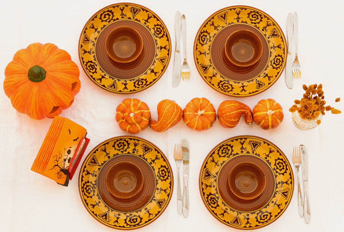 A table setting featuring four plates with bowls on top in brown and yellow color with tribal art and cutlery on the sides, placed on a white cloth with pumpkins, dried flowers and napkins in a holder.
