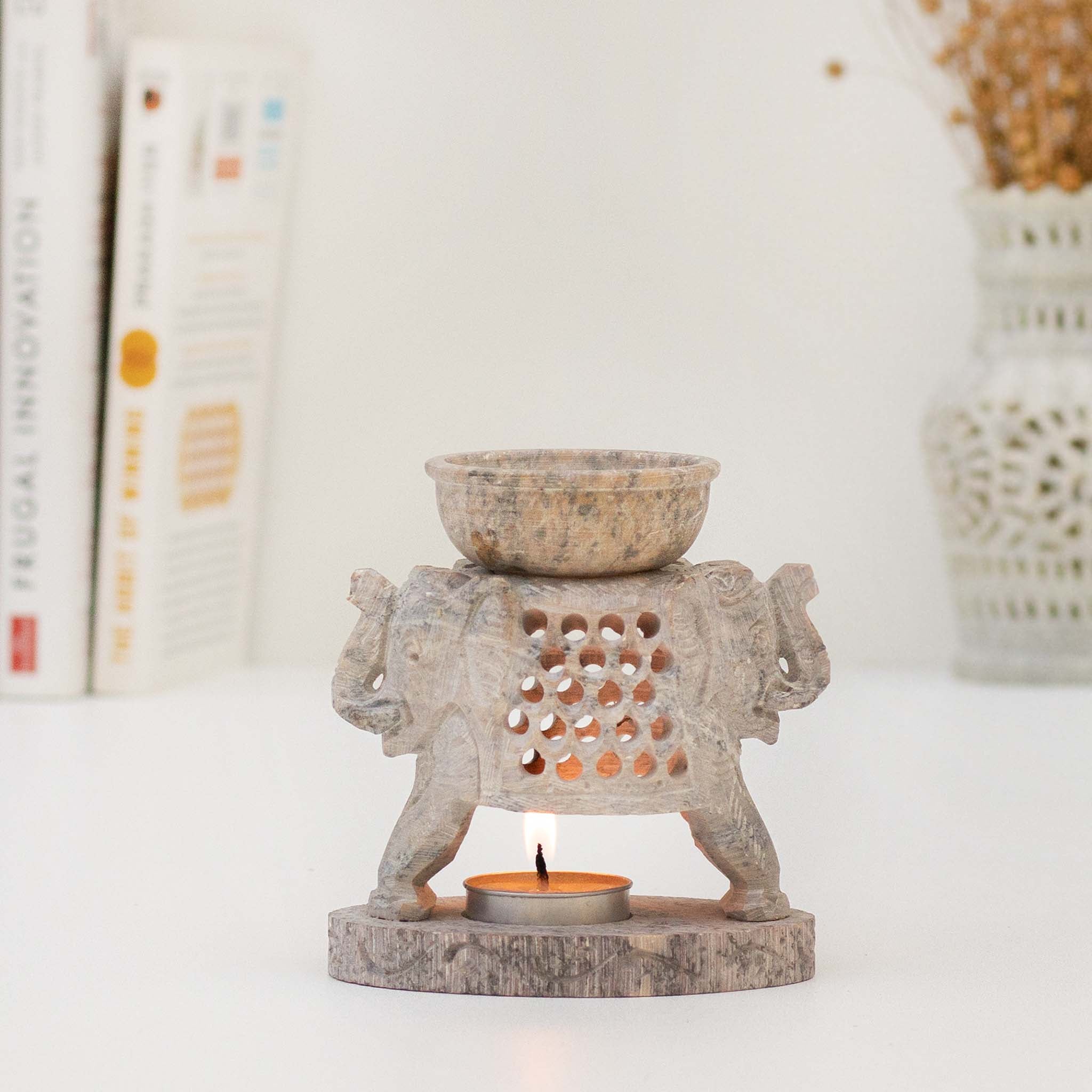 Soapstone diffuser featuring two elephant heads placed on a white surface with books, a soapstone vase of dried oats flowers and a white wall in the background.